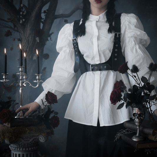 A gothic-style outfit featuring a white long-sleeve blouse with puffed sleeves, a high collar, and a black harness-style vest with silver-tone metal grommets and straps, worn by a model in a moody, dark-themed setting with candles, roses, and a skull vase.