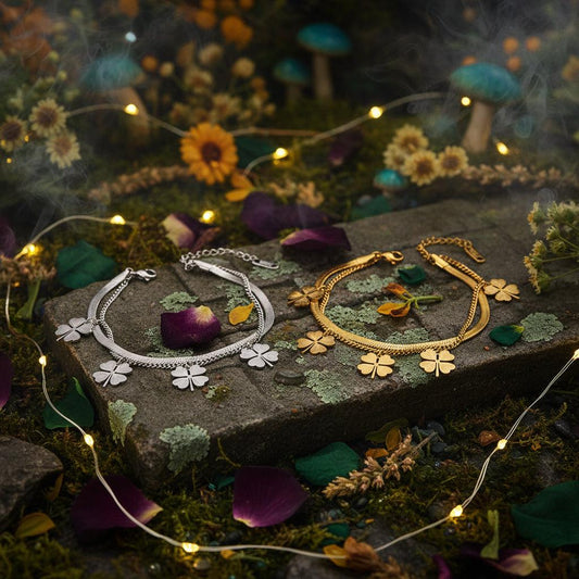 Two chain bracelets with four-leaf clover charms, one silver-toned and one gold-toned, displayed on a white card over a brown surface with warm ambient lighting.