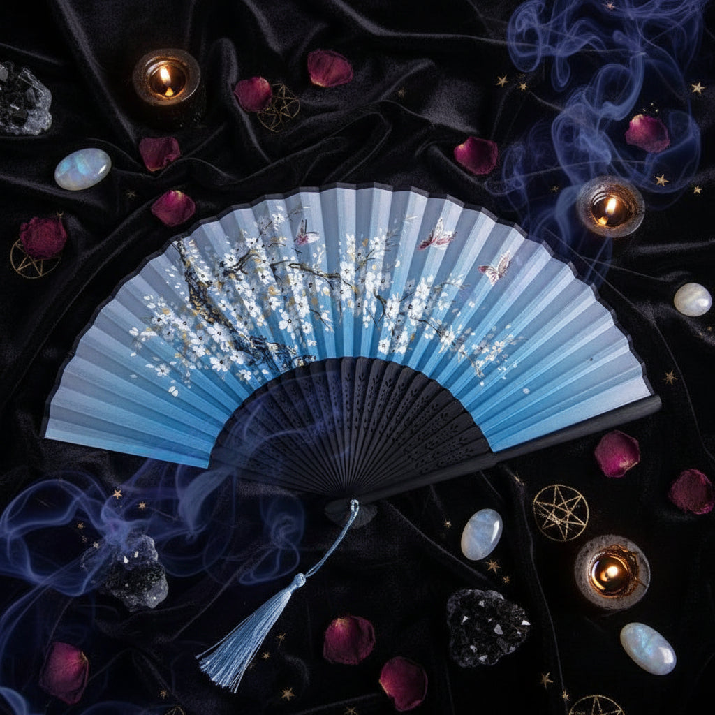 A traditional folding hand fan with a blue and black gradient design, decorated with white cherry blossoms and small butterflies. The fan is displayed on a white background with pink flowers as decorative props.