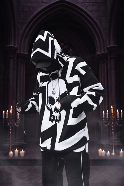 A person wearing a black and white zip-up hoodie with a large skull graphic on the front, styled with a black cap, black gloves, and a silver pendant necklace, standing against a concrete wall.