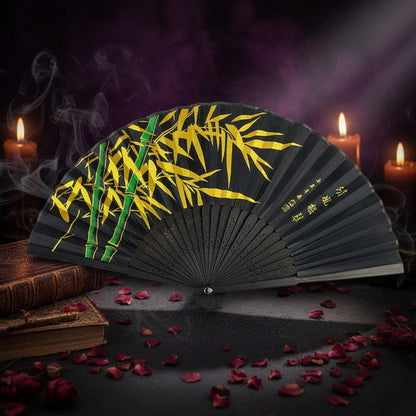 A black folding hand fan with yellow bamboo leaf patterns and green bamboo stalks, displayed on a white tray with pink flowers against a dark gray background.