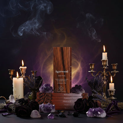 A boxed fragrance product labeled 'Agarwood' stands centered on stacked books, surrounded by lit white candles, brass candelabras, purple dried flowers, black roses, crystals, and smoky purple and gold atmospheric effects against a dark background.