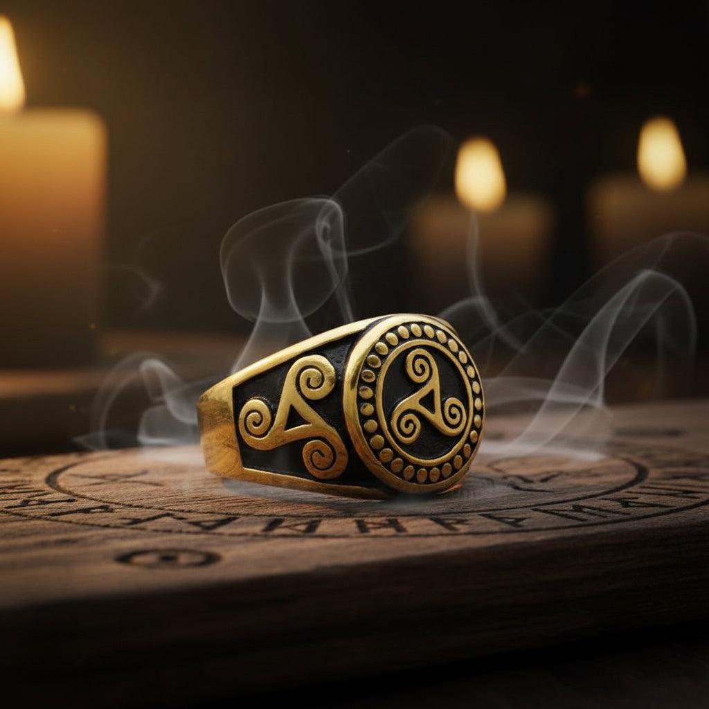 A gold-toned signet ring with black recessed detailing, featuring a Celtic triquetra symbol on one side and a circular triquetra motif on the other, resting on a wooden surface.