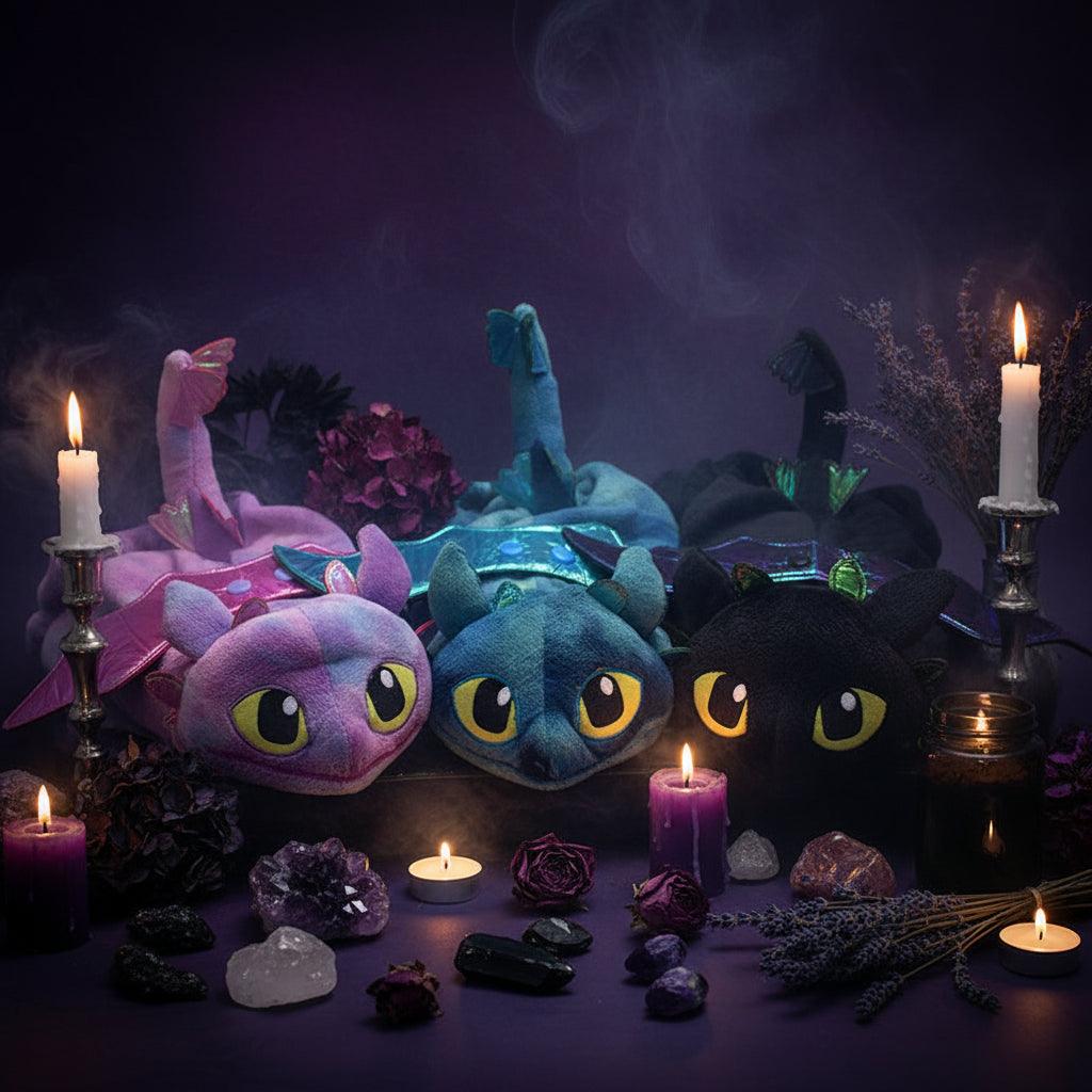Three plush dragon-shaped headbands or hair accessories displayed side by side on a plain white background. The left one is pink and purple with a long neck, the middle one is teal and blue with wings, and the right one is black with wings and large yellow eyes.