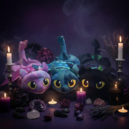 Three plush dragon-shaped headbands or hair accessories displayed side by side on a plain white background. The left one is pink and purple with a long neck, the middle one is teal and blue with wings, and the right one is black with wings and large yellow eyes.
