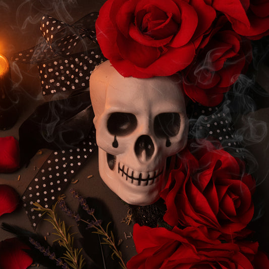 A decorative skull centerpiece with black teardrop eye makeup, surrounded by large red roses and black ribbon with white polka dots on a dark gray background.