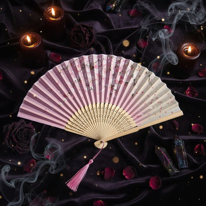 A traditional folding hand fan with pink and cream floral printed panels, a wooden handle, and a pink tassel, styled with pink flowers on a white background.