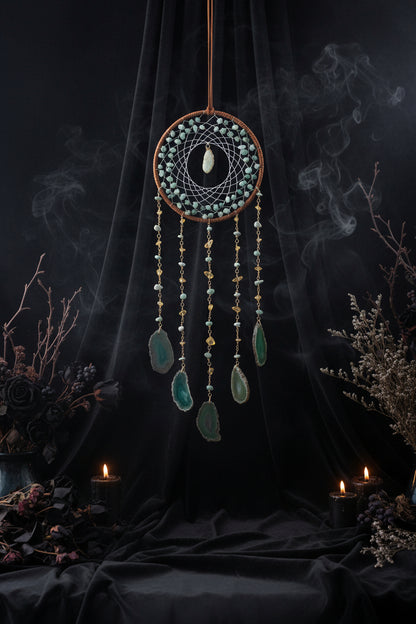 Decorative dreamcatcher with green stones and beads on a gray wall with flowers.