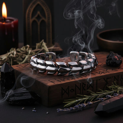 A close-up of a dark metallic link chain bracelet draped over a dark wooden box with engraved lettering, photographed against a dark background.