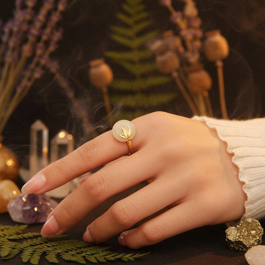 A close-up of a hand wearing a gold ring with a round pale green jadeite stone and a leaf design, resting on a wooden surface.