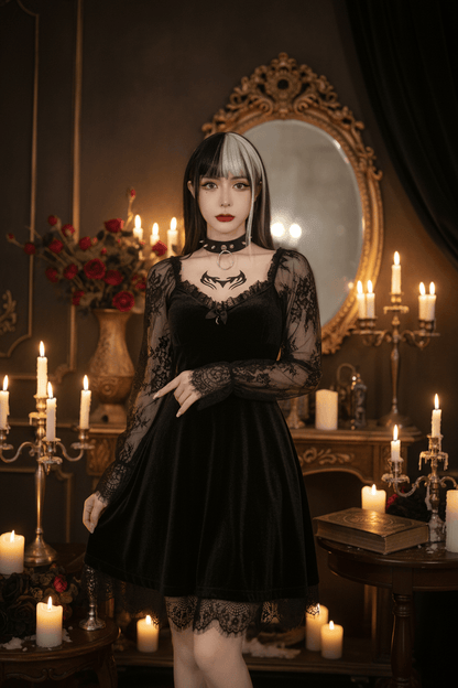 A woman wearing a black gothic dress with sheer long sleeves and a lace-trimmed bodice, standing indoors against a white wall beside white furniture and decorative statues.
