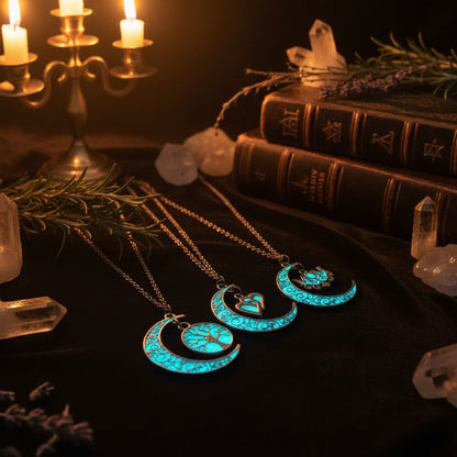 Three glowing blue crescent moon necklaces with intricate filigree patterns, each featuring a different pendant design (a tree, a heart with a bird, and a flower) on dark chains against a black background.