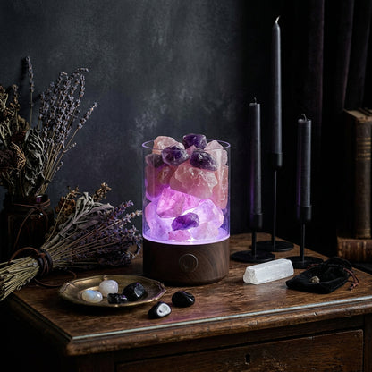 A glowing pink salt lamp in a clear cylindrical glass container with a white base, placed on a wooden surface against a dark blue background. A small vase with dried stems sits to the left.