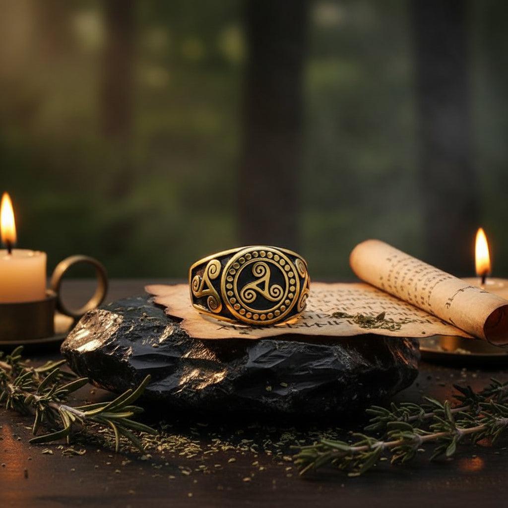 A wide band ring with a black and gold Norse-style design featuring a central triquetra symbol and ornate scrollwork, resting on a wooden surface.