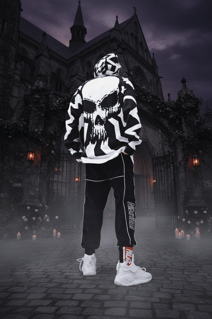A person standing with their back to the camera wearing a black and white patterned hoodie with a large skull graphic on the back, black pants with white piping and side text, and white sneakers, set against a concrete wall.