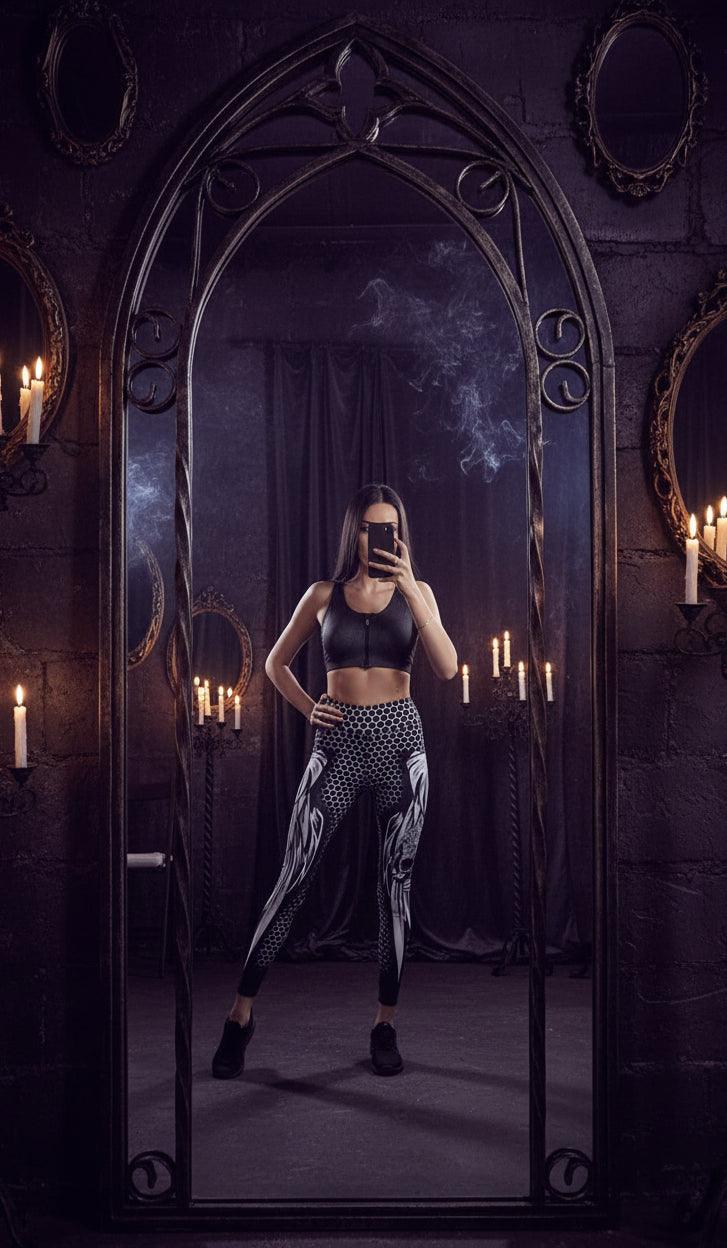 A woman takes a mirror selfie in a room showing a matching black and white athletic set, consisting of a sleeveless crop top with a front zipper and high-waisted leggings with a honeycomb pattern and white wing designs.