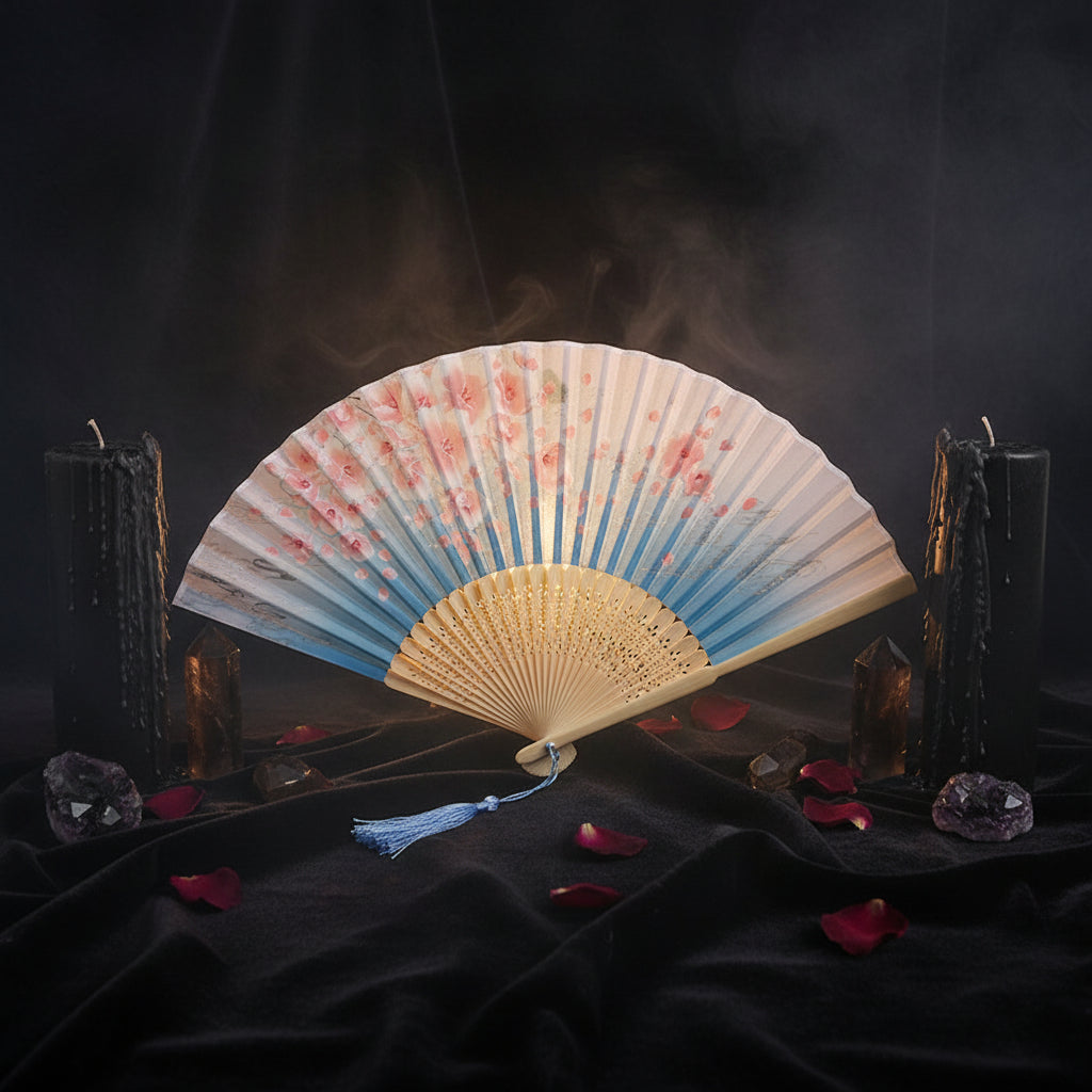 A traditional folding hand fan with a blue and white cherry blossom print, beige wooden ribs, and a blue tassel, displayed on a white fabric background with pink cherry blossom props.