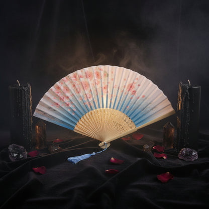 A traditional folding hand fan with a blue and white cherry blossom print, beige wooden ribs, and a blue tassel, displayed on a white fabric background with pink cherry blossom props.