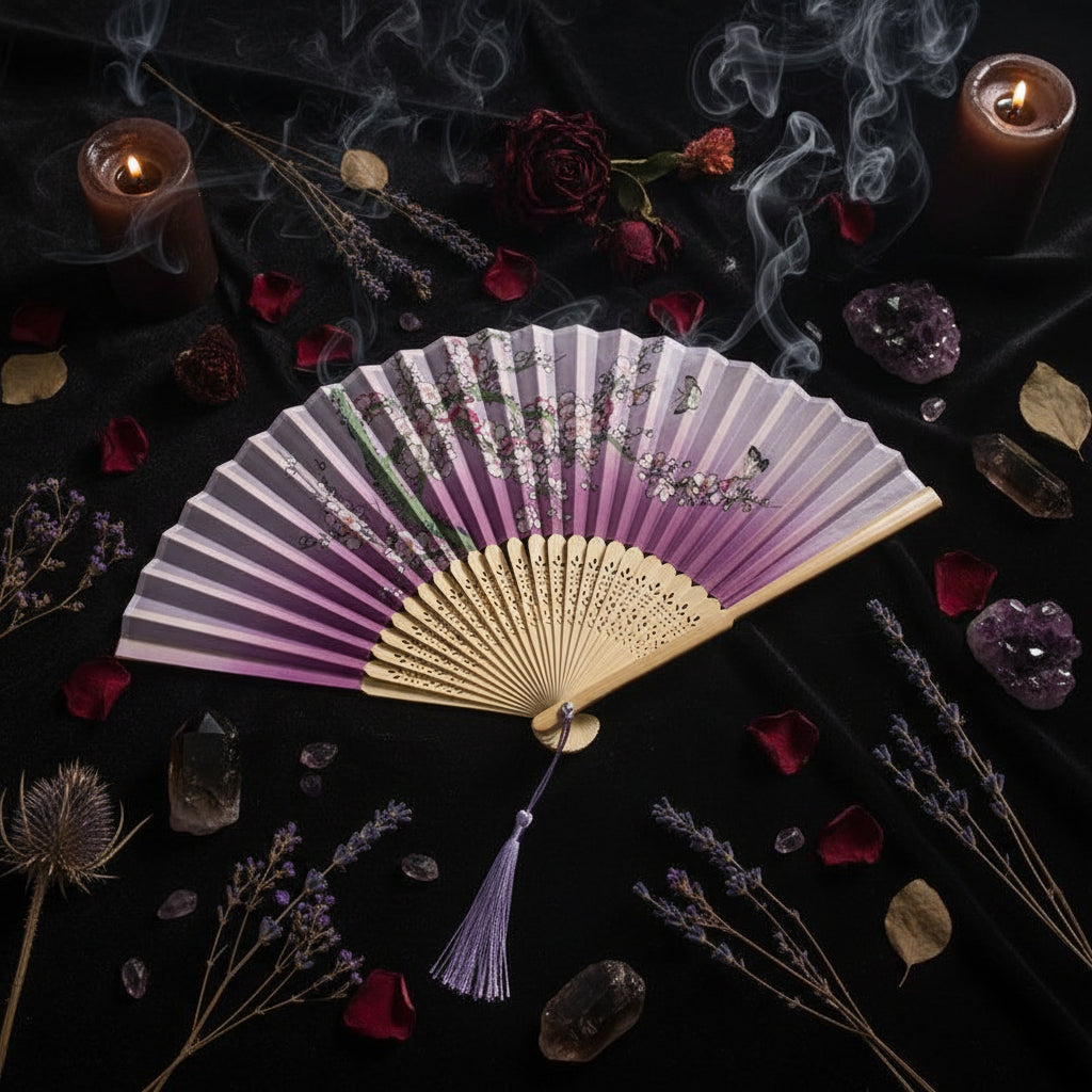A traditional folding hand fan with a pink and purple gradient design, decorated with cherry blossom branches and butterflies. The fan features a beige bamboo core and a purple tassel, styled with pink flowers on a white background.