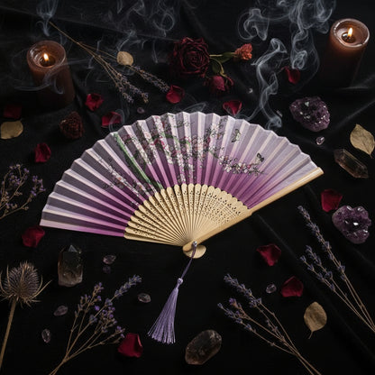 A traditional folding hand fan with a pink and purple gradient design, decorated with cherry blossom branches and butterflies. The fan features a beige bamboo core and a purple tassel, styled with pink flowers on a white background.