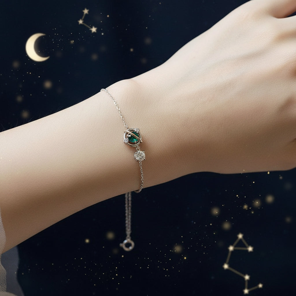 A delicate silver bracelet with a small oval charm and a dangling clear stone, shown worn on a person's wrist against a soft white fabric background.