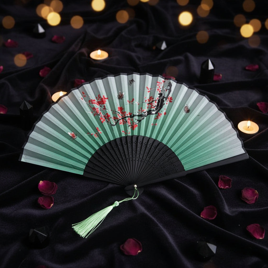 A traditional folding hand fan with a light green and white striped design, decorated with red cherry blossoms and black branches. The fan has a black frame, a green tassel, and is styled on a white background with pink cherry blossom branches.