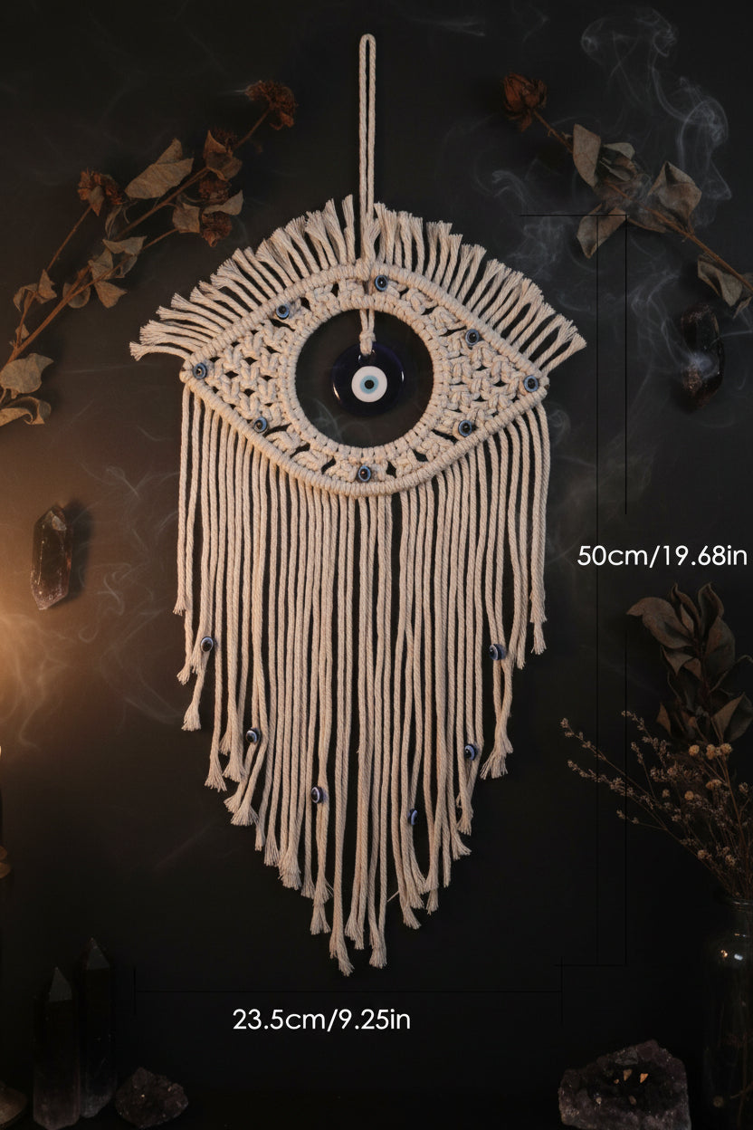 A macrame wall hanging shaped like an eye, made from off-white cord with a blue-and-white evil eye motif in the center and small blue bead accents. Measurement lines and text indicate dimensions of 50cm in height and 23.