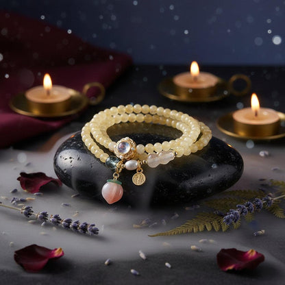 A double-strand beaded bracelet with pale yellow and cream beads, a gold-toned moonstone centerpiece, a white pearl, a blue bead, and a dangling peach-shaped pink stone charm, displayed on a gray box.