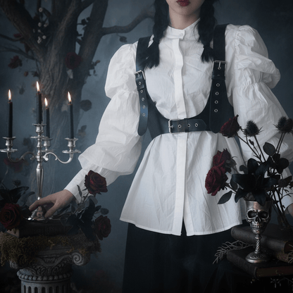 A gothic-style outfit featuring a white long-sleeve blouse with puffed sleeves, a high collar, and a black harness-style vest with silver-tone metal grommets and straps, worn by a model in a moody, dark-themed setting with candles, roses, and a skull vase.