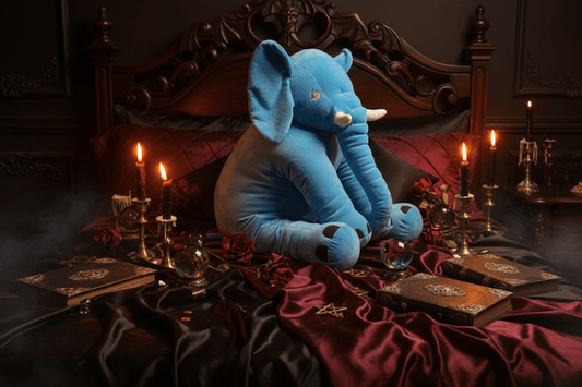 A large blue plush elephant toy sitting on a floral bedspread in a nursery-like setting with a white crib edge, framed picture, and potted flowers in the background.