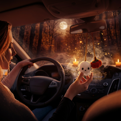 A person driving a car while holding a phone, with two small hanging ornaments below the rearview mirror: a white ghost and a red pumpkin.