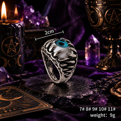 A silver-toned ring with a textured, claw-like design surrounding a white oval center and a blue evil-eye motif, shown on a white background with a 2cm size indicator and text listing styles and weight.