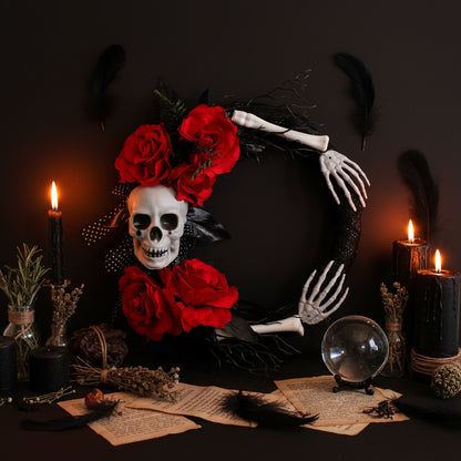 A gothic Halloween wreath decoration featuring a white skull, red roses, black mesh, skeleton hands, and black twigs on a plain white background.