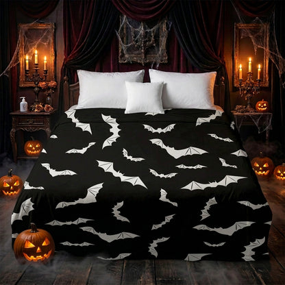 A bed dressed in a black blanket or sheet featuring an allover white bat print, styled in a bedroom with gray and white pillows, a wooden side table, and a woven pouf.