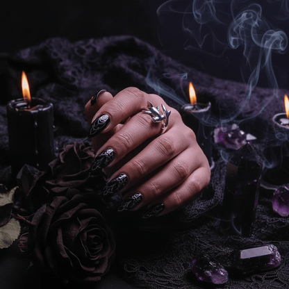 Hand with ornate nails and shark rings holding a small object, surrounded by candles and crystals on a dark background