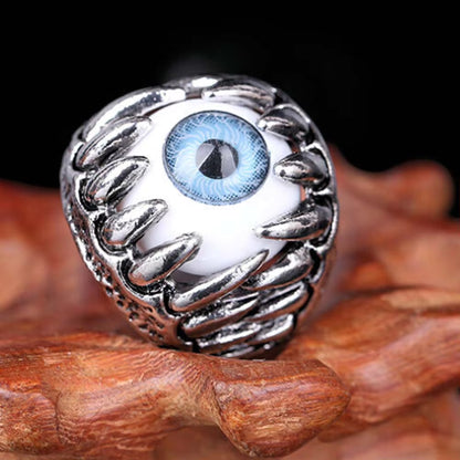 A silver-toned ring featuring a large white globe with a detailed blue and black human eye in the center, displayed on a piece of wood against a dark background.