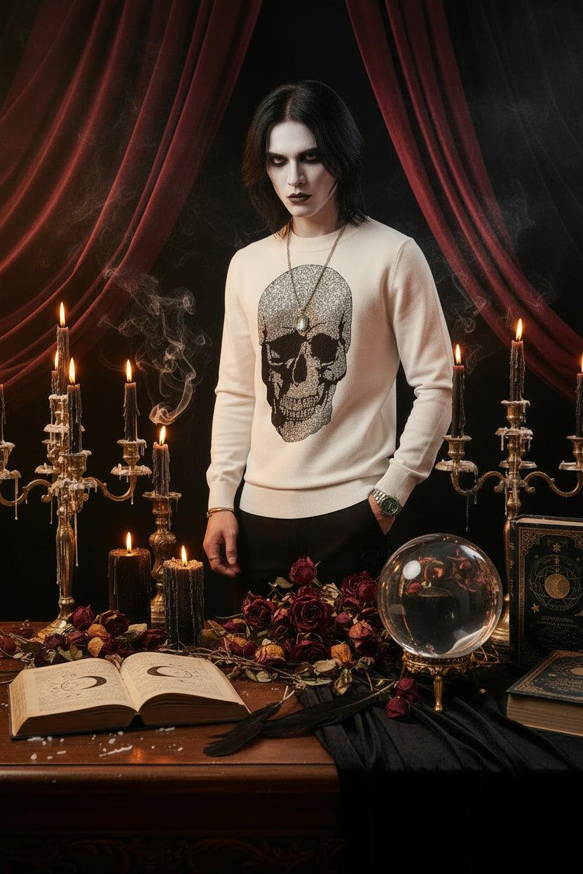 A man wearing a white long-sleeve crewneck sweater featuring a large black skull graphic embellished with sparkling rhinestones on the front, paired with a silver chain necklace.