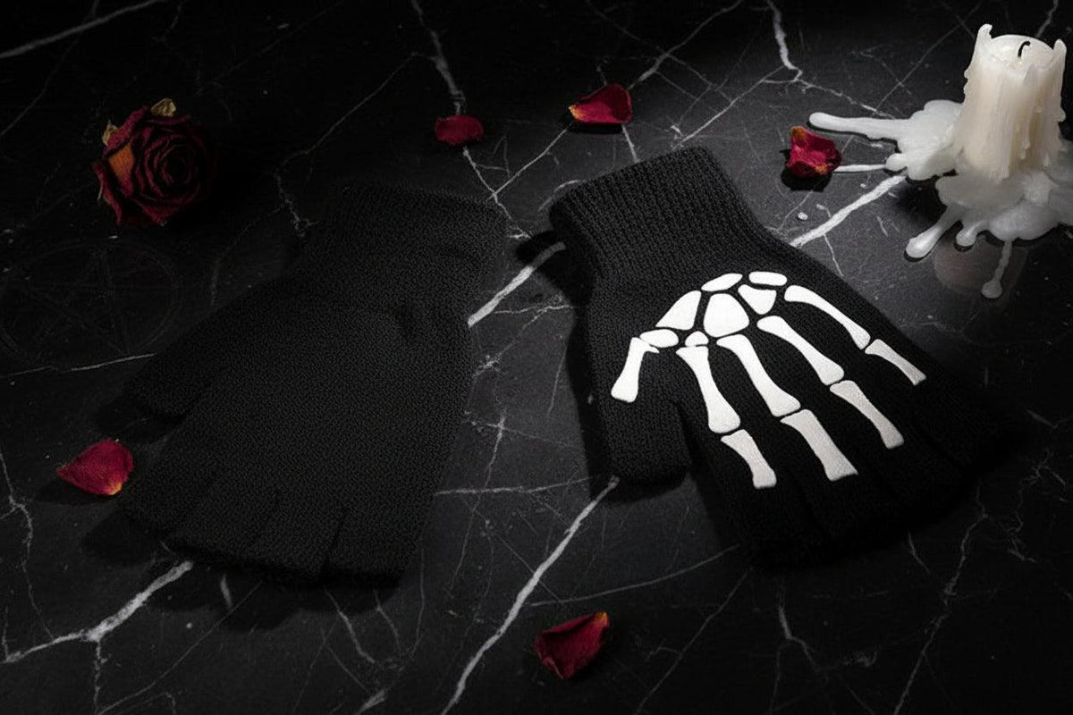 A pair of black fingerless knit gloves with a white skeleton hand graphic on one glove, displayed on a dark wood background with decorative flowers and a wooden box edge.