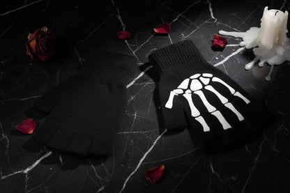 A pair of black fingerless knit gloves with a white skeleton hand graphic on one glove, displayed on a dark wood background with decorative flowers and a wooden box edge.