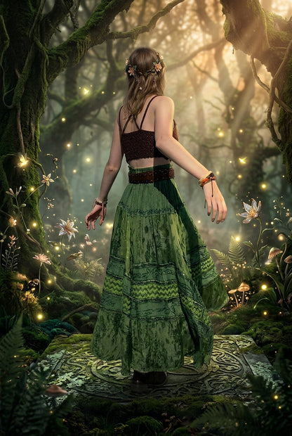 A woman wearing a green bohemian-style maxi skirt with a brown crop top stands on a rocky beach with the ocean in the background.