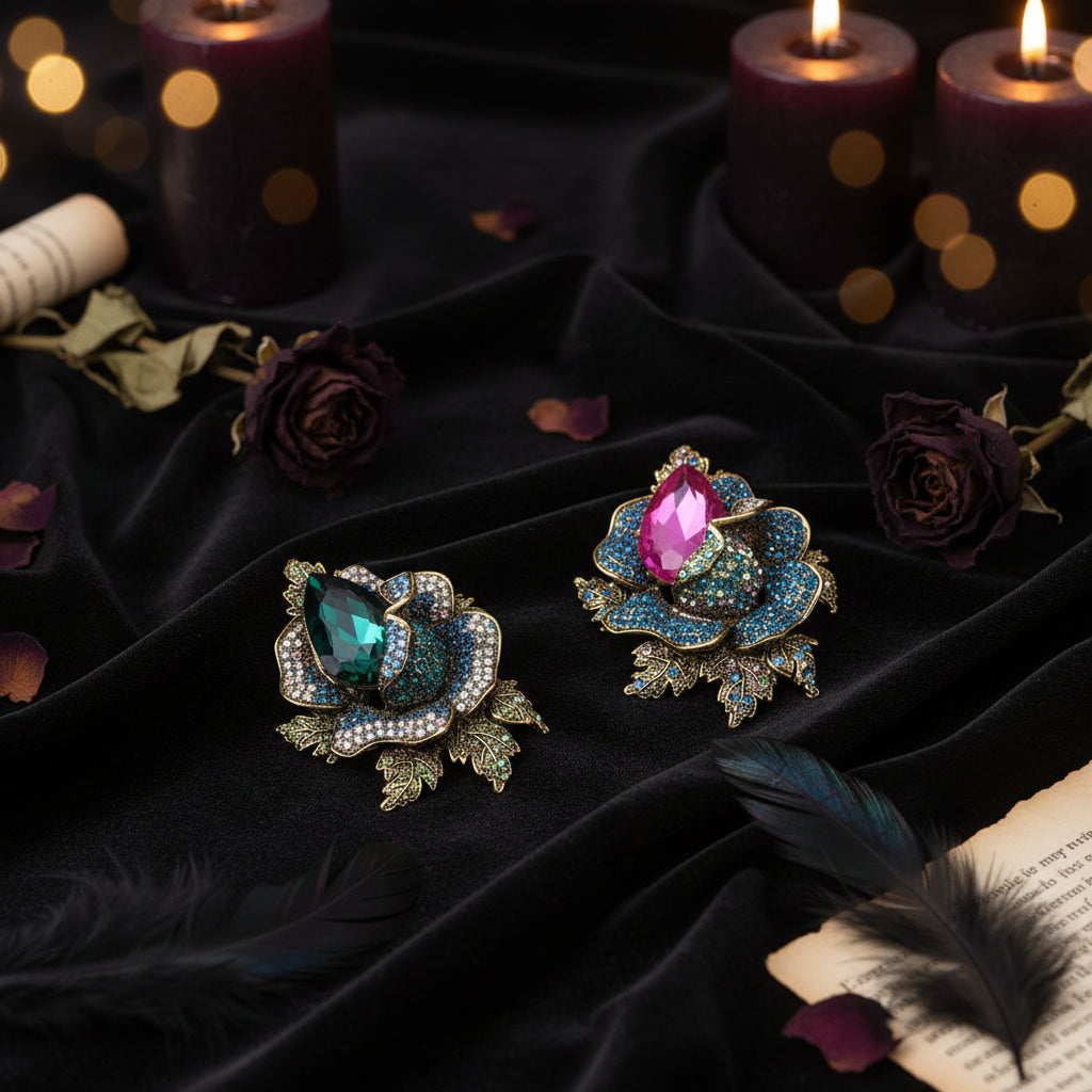 Two ornate rose-shaped brooches with large iridescent gemstones, one in emerald green and one in pink, set in gold-tone metal with blue rhinestone accents, displayed on a white grid background with decorative petals and a book corner.