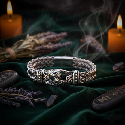 A silver-toned braided chain bracelet with an ornate central clasp featuring a Viking helmet motif, displayed on a dark textured stone surface.