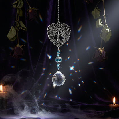 Silver filigree heart suncatcher with blue crystal beads and a clear teardrop crystal pendant, hanging against a soft green background with blurred white flowers and greenery.