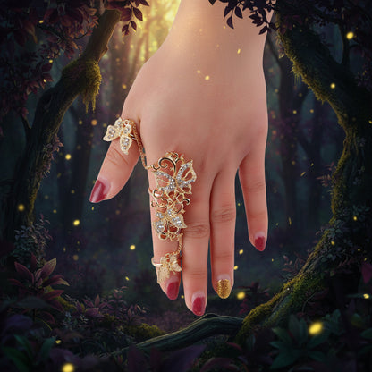 A gold-toned finger ring with butterfly and floral motifs, embellished with clear rhinestones, shown worn on a hand with red and gold manicured nails against a light gray background.