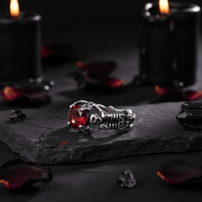 A silver-toned gothic ring featuring a detailed skeleton design and a red gemstone, displayed on a textured tan fabric background.