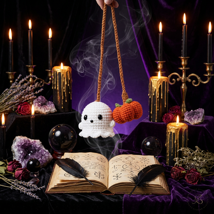 A small crocheted Halloween car accessory hanging from a rearview mirror, featuring a white ghost and an orange pumpkin with green stems, shown inside a car with a blurred house and autumn trees in the background.