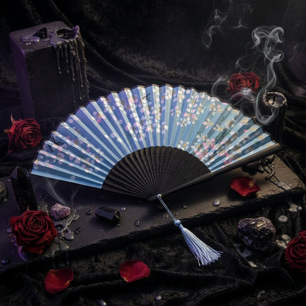 A traditional folding hand fan with a light blue floral pattern and a black frame, displayed on a white fabric background with pink cherry blossom flowers.