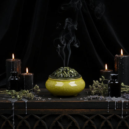 A small ceramic incense burner with a rounded body and perforated lid, shown on a wooden surface against a black background. The glaze is a pale yellow-green with a glossy finish, and the lid has a darker green floral relief design.