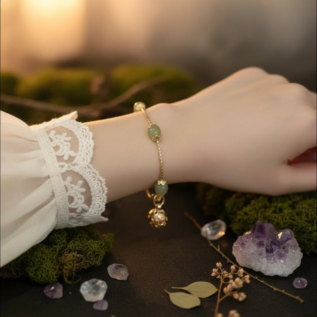 A gold chain bracelet with two green jade-like beads and a dangling gold charm, shown worn on a person's wrist.