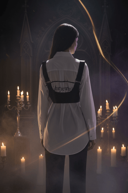 A black faux-leather vest with wide shoulder straps and a wide waistband, shown on a model from the back wearing a white long-sleeve shirt with black text.